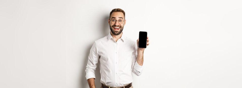 Amazed handsome businessman showing smartphone screen app, standing over white background.