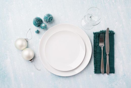 Christmas table setting with white dishware, silverware and green decorations on blue background. Top view, flat lay
