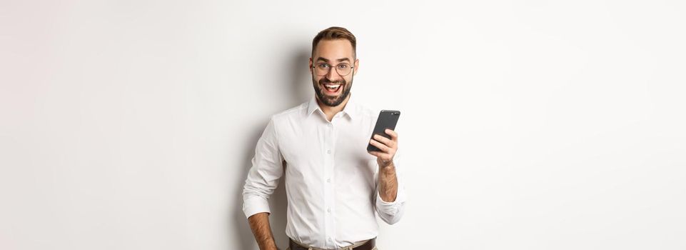 Excited business man using mobile phone, looking amazed, standing over white background.