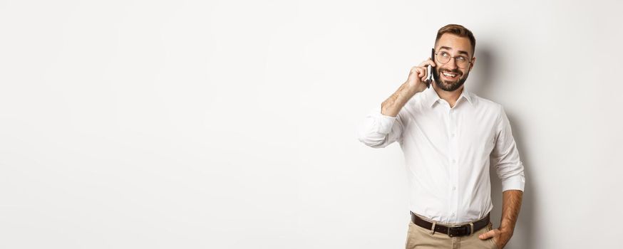 Successful business man in glasses talking on mobile phone, looking satisfied and smiling, standing against white background.