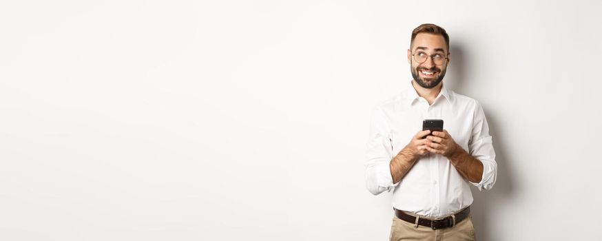 Thouthful handsome manager using mobile phone and thinking about answering message, looking at upper left corner and smiling, standing over white background.