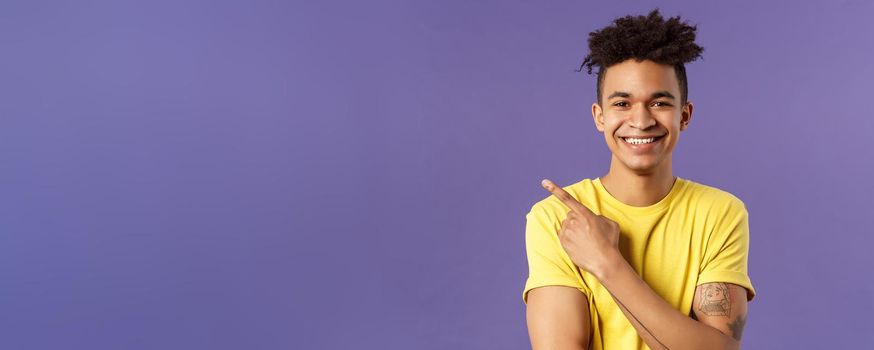 Close-up portrait of enthusiastic, happy young hipster male with dreads, beaming smile and pointing finger upper left corner, present cool product, introduce something really good, purple background.