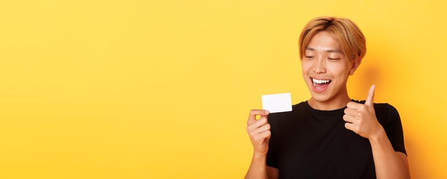 Close-up of happy and satisfied asian handsome guy, showing credit card and thumbs-up in approval, smiling amazed, standing yellow background.