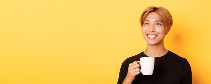 Close-up of happy handsome young asian guy with blond hair, looking dreamy and smiling while drinking coffee or tea, standing over yellow background.