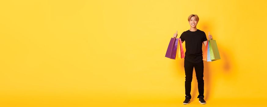 Full-length of attractive korean guy in black clothes, showing thumbs-up, holding shopping bags, yellow background.