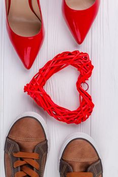 Vertical shot top view shoes and heart. Red womens shoes and brown mens footwear on white desk.