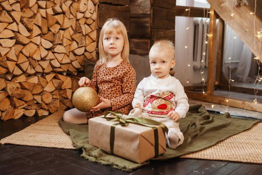 Little brother and sister play on Christmas eve in a beautiful house decorated for the New Year holidays. Children are playing with a Christmas gift. Scandinavian-style interior with live fir trees and a wooden staircase.