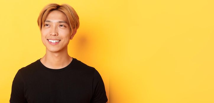 Close-up of handsome stylish korean guy with fair hair, looking dreamy and hopeful upper left corner, smiling pleased, standing yellow background.