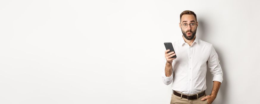 Surprised handsome man reading interesting info online, holding smartphone, standing over white background.