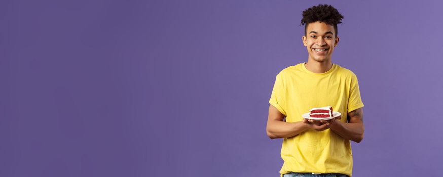 Celebration, party and holidays concept. Portrait of handsome young smiling man, feeling happy likes eating desserts, ordered delivery from local cafe to have some delicious cake, purple background.