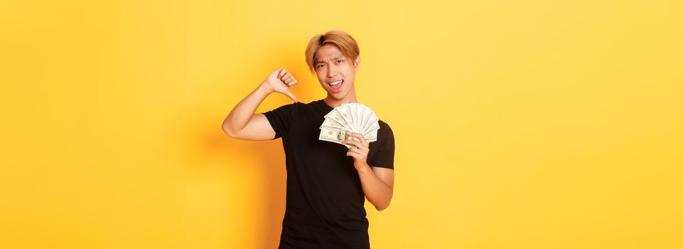 Sassy handsome asian guy pointing finger at cash and looking pleased. Korean man borrowed money, standing yellow background.