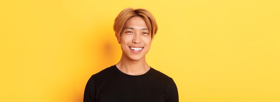 Close-up of handsome stylish asian guy with fair hair, smiling happy over yellow background.