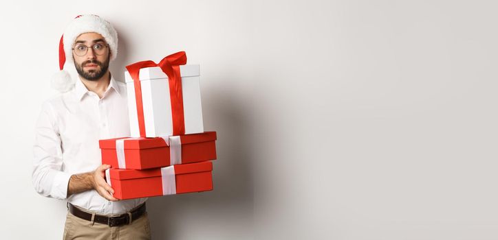 Merry christmas, holidays concept. Confused man in Santa hat holding pile of presents, found gifts under xmas tree, standing against white background.