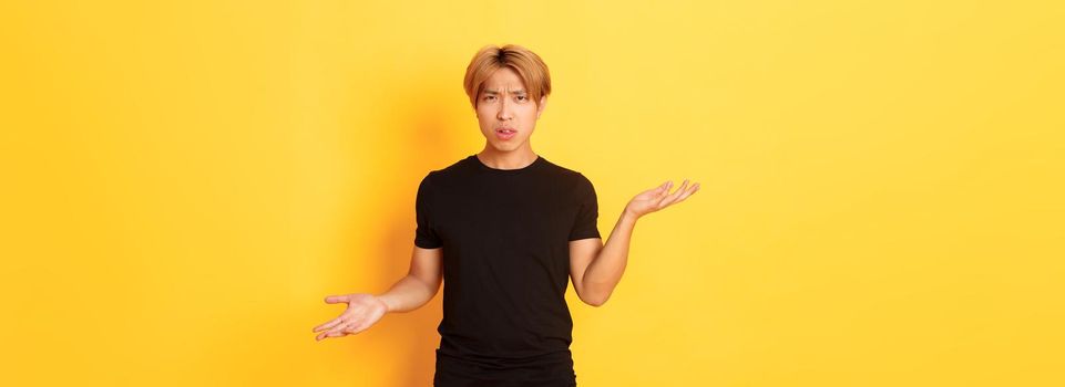 Portrait of confused and annoyed asian guy spread hands sideways, cant understand something, standing yellow background.