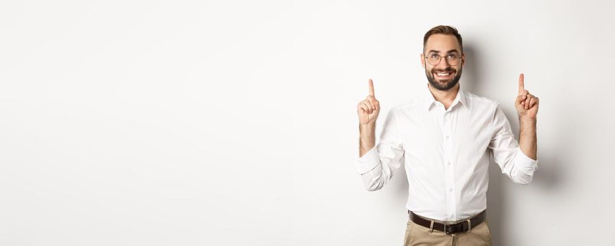 Confident handsome businessman showing logo, pointing and looking up at announcement, standing over white background.