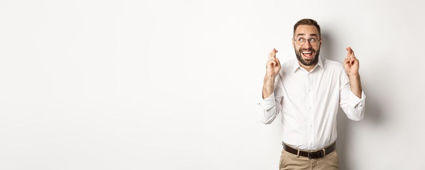 Excited and hopeful businessman making a wish, cross fingers and waiting, standing over white background.