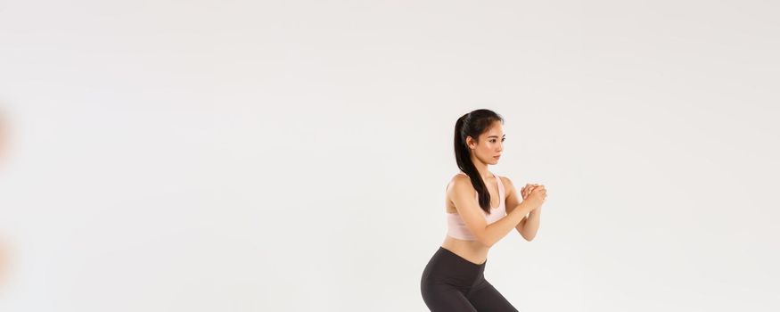 Full length side view of focused slim asian girl doing fitness training, female athlete clasp hands together and perform squats exercises with stretching resistance band, workout equipment.