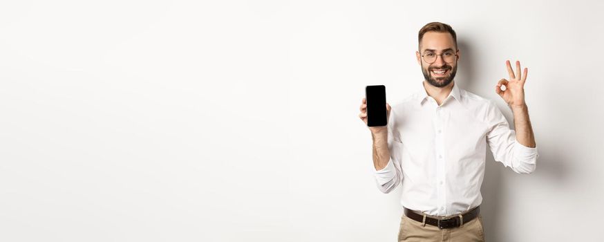 Satisfied young manager showing smartphone screen and okay sign, recommending application, standing over white background.