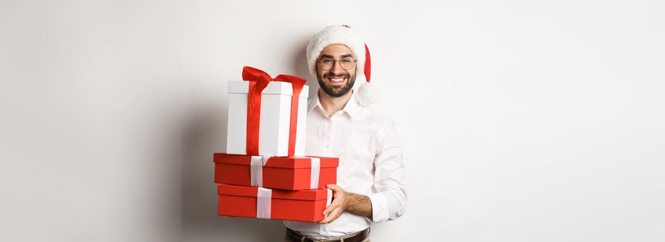 Merry christmas, holidays concept. Happy young man smiling, holding gifts in boxes and wearing santa hat, white background.