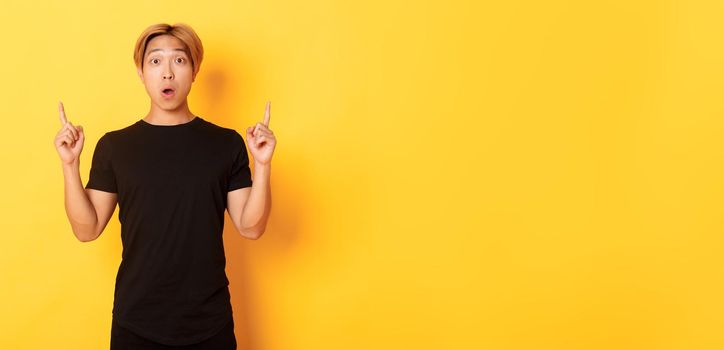 Portrait of astonished asian guy in black t-shirt, open mouth fascinated, pointing fingers up, yellow background.