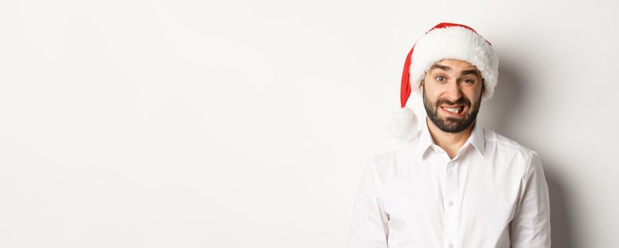 Close-up of awkward guy in santa hat apologizing, feeling uncomfortable, standing over white background. Christmas concept.