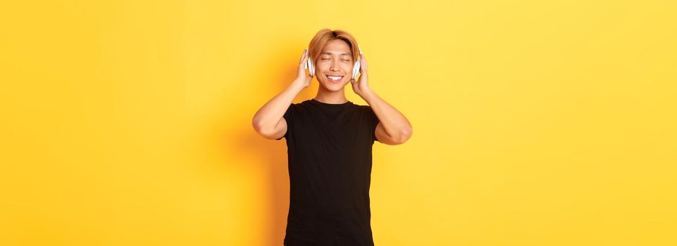 Relaxed and pleased handsome asian guy close eyes from satisfaction while listening music in wireless headphones, standing yellow background.