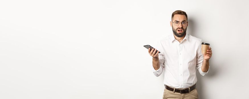 Image of man drinking coffee, feeling confused about strange message on mobile phone, standing over white background.