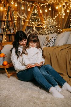 A little girl with her mother in a cozy home environment on the sofa next to the Christmas tree. The theme of New Year holidays and festive interior with garlands and light bulbs
