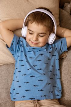 Little caucasian boy with blue shirt and white headphones listening music or audio book on the couch at home. Cute child relaxing, resting in his room