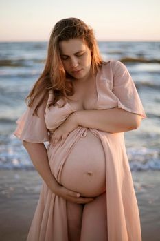 Young pregnant woman with a beautiful sea view on the background. Happy and calm pregnant woman with long hair and pink dress standig on the beach. Romantic view, ocean, sunset, maternity.