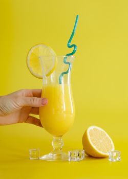 Fresh orange juice in glass, decorated with slice of lemon and blue straw. Summer cocktail on the yellow background. Colorful design