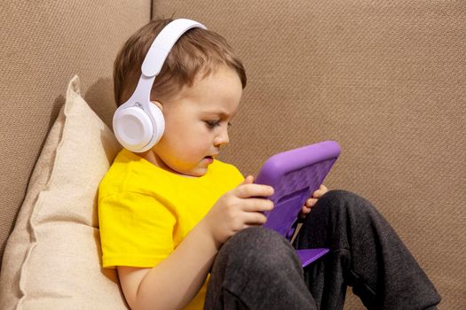 Little caucasian boy with yellow shirt playing game on digital tablet at home. Portrait of a child at home watching cartoon on violet tablet. Modern kid and education technology