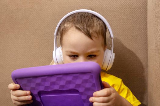 Little caucasian boy with yellow shirt playing game on digital tablet at home. Portrait of a child at home watching cartoon on violet tablet. Modern kid and education technology