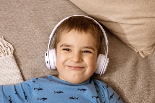 Little caucasian boy with blue shirt and white headphones listening music or audio book on the couch at home. Cute child relaxing, resting in his room. Smiling, happy kid
