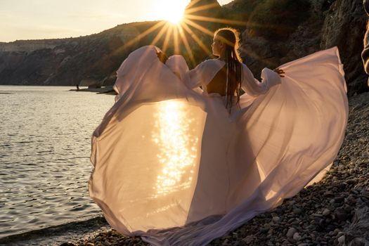 A mysterious female silhouette with long braids stands on the sea beach with mountain views, Sunset rays shine on a woman. Throws up a long white dress, a divine sunset