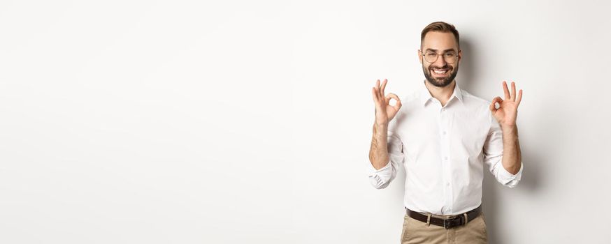 Satisfied handsome businessman showing ok sign, gurantee quallity, standing pleased against white background.