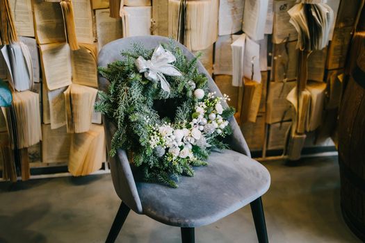 Christmas wreath made of natural coniferous branches and decorated with decorative white elements. A beautiful Christmas wreath is lying on a velour grey chair.