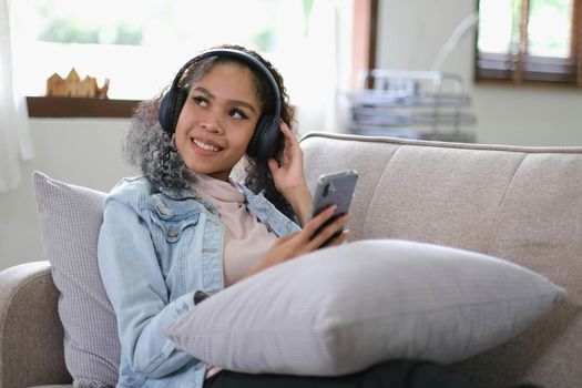 Young relaxed woman listening to the music resting on the sofa at home. Caucasian girl using smart phone and headphones for music application online..