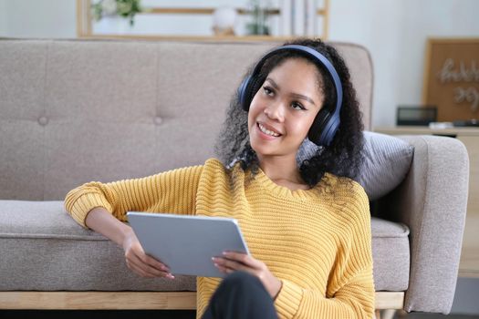 Photo of beautiful dark skin wavy lady homey mood holding digital tablet earflaps listening new audio sitting floor near couch casual clothes yellow at