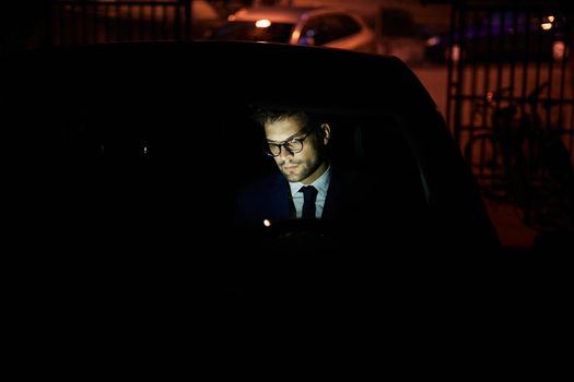 Portrait of a young businessman with tablet or laptop working late at night in office or looking at a device screen in car