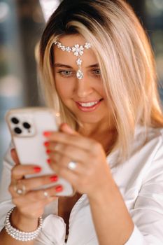 A positive young woman in a white dress and an ornament on her head was smiling, holding a phone in her hands and looking into it.