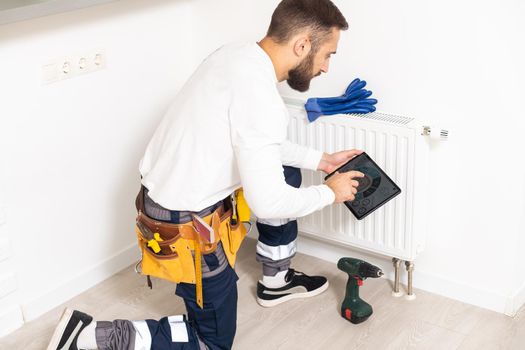 Man in work overalls using wrench while installing heating radiator in room. Young plumber installing heating system in apartment. Concept of radiator installation, plumbing works and home renovation