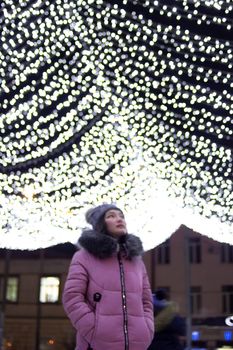Young woman looks up, central downtown with christmas lights at night, sky festive illumination shine and glow, urban holiday, vertical defocus image with copy space, low angle