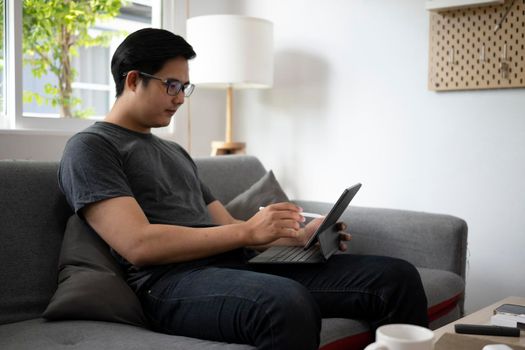 Handsome asian man resting on couch and browsing internet with computer tablet.