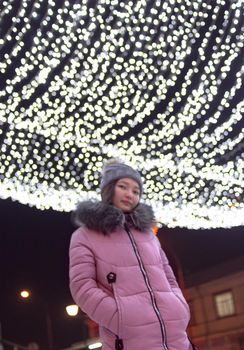 Young woman on central downtown with christmas lights at night, sky festive illumination shine and glow, urban holiday, vertical defocus image with dynamic slanted horizon, low angle