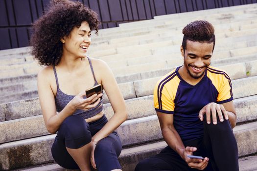 athletic latin woman smiles happy with a smiling black sports man and have fun with mobile phone on the stairs during a rest in the workout, technology in sport and healthy lifestyle concept