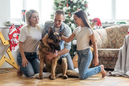 Friendly family is playing with dog near Christmas tree. They are sitting and laughing.