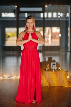 Portrait of a smiling girl in a red dress stands in the interior with her hands clasped in front of her.