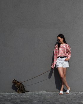A young woman walks with a gray tabby cat on a leash against a gray wall