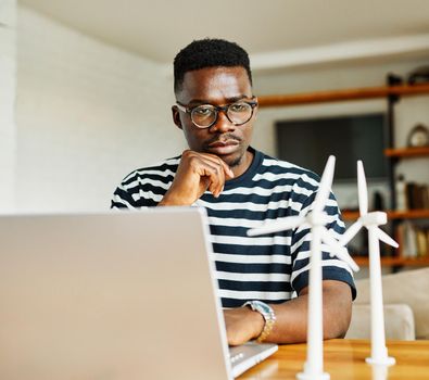 Portrait of a young black man using laptop and looking at windmill models at home